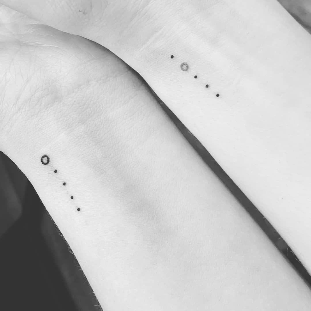 Two wrists with minimalist tattoos of dots and a small circle in a straight line
